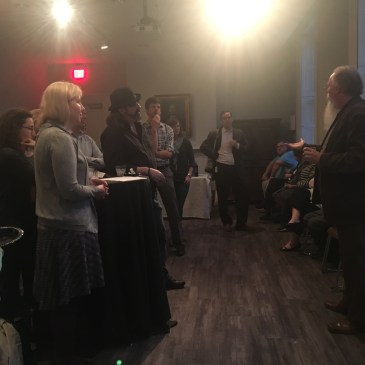 Speaker in front of a small room of people with cocktails.