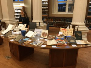 Greeter behind curved table with a number of materials, like a teapot and books, spread out on it.