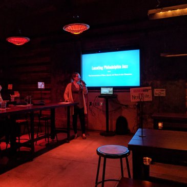 Chelsea Reed speaking to the audience with a slide on the screen behind her that says "Locating Philadelphia Jazz"