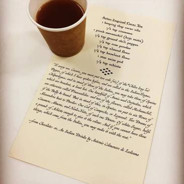 Coffee cup resting on drink recipe for Aztec-Inspired Cacao Tea printed on parchment looking paper in decorative print.